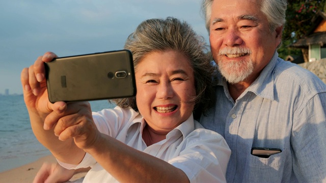资深亚洲夫妇使用智能手机自拍在海滩上与幸福的情感在一起。有生活方式、退休和科技观念的人。