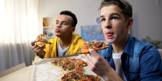 两个多民族男性青少年在吃披萨看电视，闲暇时间