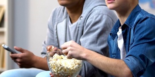 兴奋的青少年吃着爆米花，看着有线电视上紧张的犯罪剧