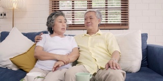 一对亚洲老夫妇在家里客厅的沙发上放松时，开心地微笑着，看着镜头。享受时光的生活方式高级家庭在家里的概念。看着相机的人像。