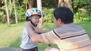 爸爸把头盔戴在亚洲2 - 3岁蹒跚学步的小男孩身上，爸爸和儿子玩平衡自行车视频素材模板下载