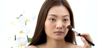 亚洲美女用腮红刷在脸颊上化妆成与蝴蝶兰一样美丽的白色背景。人以美容、保健、情感为理念。