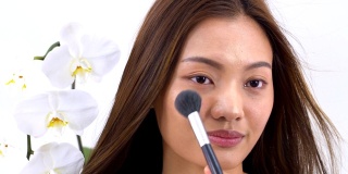 大头照:亚洲美女用腮红刷化妆，美丽的蝴蝶兰在白色的背景。人以美容、保健、情感为理念。