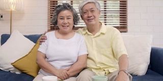 一对亚洲老夫妇在家里客厅的沙发上放松时，开心地微笑着，看着镜头。享受时光的生活方式高级家庭在家里的概念。看着相机的人像。