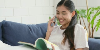 年轻美丽的亚洲女人谈论智能手机与幸福坐在沙发上看书，人在电话，生活方式
