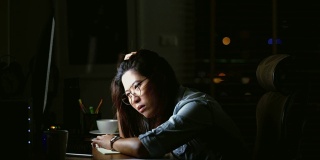 迷人的亚洲女人工作到很晚，思考与严肃的行动在桌子前的电脑显示器桌面在黑暗的工作场所，工作到很晚和努力的概念