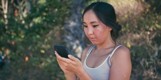 年轻快乐美丽的亚洲女人在户外使用手机在自然运动服装