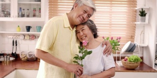 亚洲老夫妇感到幸福的微笑，拿着花，看着镜头，而放松在厨房在家里。生活方式老年人家庭享受居家时光的概念。看着相机的人像。