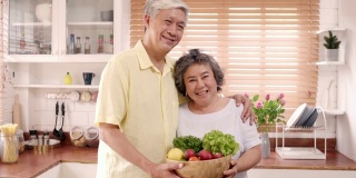 亚洲老夫妇感到幸福的微笑，拿着水果，看着镜头，而放松在厨房在家里。生活方式老年人家庭享受居家时光的概念。看着相机的人像。