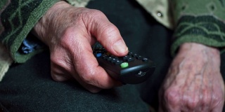一个老年人看电视，娱乐，一个奶奶的手满皱纹使用遥控器的特写。