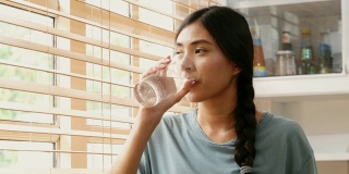 特写年轻的亚洲妇女在厨房里喝水在家，人们健康的生活方式