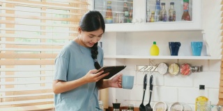 年轻美丽的亚洲女人在家里的厨房里一边喝着咖啡一边用着数字平板电脑，人和科技，Z一代的生活方式