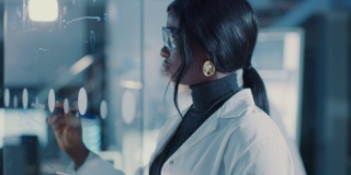 在研究实验室聪明美丽的非裔美国女科学家穿着白大褂和防护眼镜在玻璃白板上写公式，参考她的平板电脑
