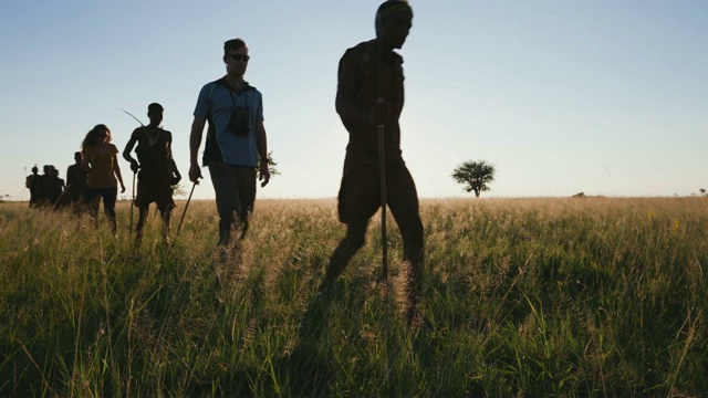 博茨瓦纳，一群游客在Makgadikgadi草原上与身穿传统服装的桑族人/布希曼人一起在丛林中漫步，身后是夕阳