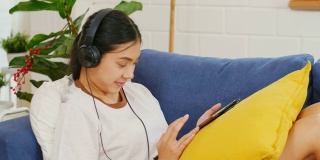 年轻美丽的亚洲女人坐在家里客厅的沙发上，用数码平板电脑和耳机听音乐，人和科技，Z一代的生活方式