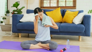 年轻的亚洲女子运动后喝水，坐在自家客厅的运动垫上背景，人们健康的生活方式，幸福视频素材模板下载