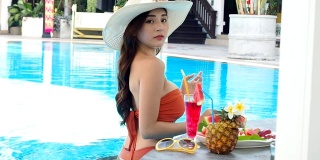 夏天的概念。性感的亚洲女人在橙色比基尼与热带饮料在游泳池。