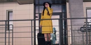 穿着黄色雨衣，戴着帽子，一头乌黑长发的漂亮女人走到酒店的门廊上，摘下眼镜，环顾四周