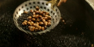 亚洲男人的手把栗子放在锅里，放在漏勺上。(特写)在泰国，用大平底锅烤栗子和咖啡豆。