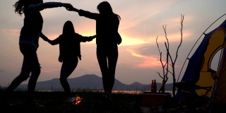低角度的观点:朋友跳舞围绕篝火在夏天，而露营附近的湖在日落
