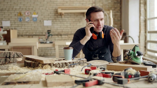 轻松的家具制造商在工作场所打电话。他面前的工作台上放着木材、工作工具、一次性咖啡杯和木屑