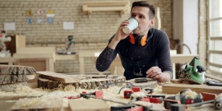 追踪中等镜头的专业细木工喝咖啡和浏览智能手机站在他的木工车间的工作台