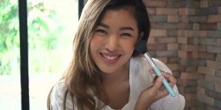 年轻的亚洲微笑女人使用化妆刷和看着相机在家里。