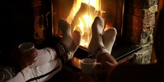 年轻夫妇在壁炉旁用圣诞装饰的袜子暖脚，喝着茶。温馨圣诞之家概念4K镜头