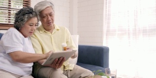亚洲老年夫妇在客厅用平板电脑搜索药物信息，夫妇在家里躺在沙发上放松时一起使用时间。老年人家庭健康在家的概念。