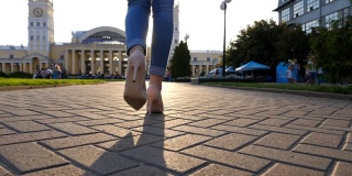 跟着女性的脚高跟鞋走在城市的街道上。穿着高跟鞋进城的年轻女子的脚。女孩踩在人行道上，太阳耀斑在背景。后视图特写
