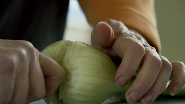 一位年长的白人妇女从盛满蔬菜的金属碗里拿出茴香，然后用菜刀将其切在切菜板上