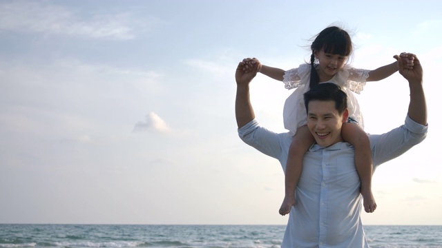 快乐的爸爸和女儿在蓝天背景下的海滩上玩耍。缓慢的运动。幸福的家庭的概念。