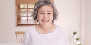 亚洲老年妇女感到幸福的微笑和看着镜头，而放松在客厅的沙发在家里。生活方式高级女性在家的概念。