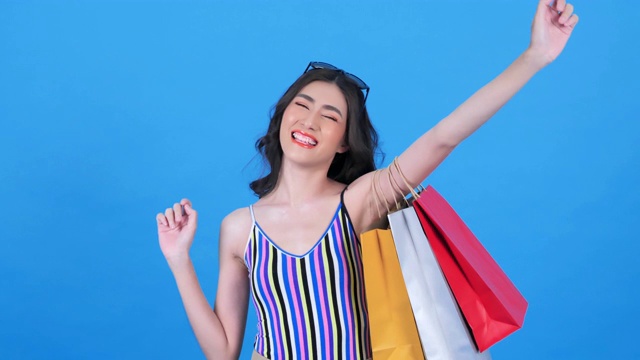 一个兴奋和微笑的年轻亚洲妇女与购物袋的肖像。女人在夏天购物，喜欢购物袋孤立在蓝色的背景。消费主义、购物、生活理念