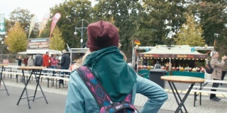 慢动作年轻的白人女游客穿着蓝色夹克，背着背包在城市集市上散步。背景是不知名的商店和人