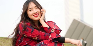 美丽的中国女孩穿着红衣服坐在草地上，享受微风吹拂她的长发，看着镜头，甜美的微笑，4k电影，慢镜头。