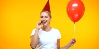 欢快的年轻女士戴着派对帽吹号角，拿着红色气球，过生日