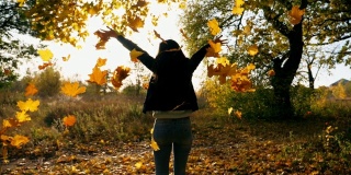 一个陌生的年轻女子在秋天的公园里跳着，扔着黄色的枫叶。彩叶飘落。女孩举起手，享受这一刻。美丽的自然景观。慢动作