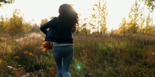年轻女子跑过秋天的森林手里拿着黄叶的花束并把它们扔出去。女士在五彩缤纷的秋天森林里玩耍。日落时分，女孩在户外慢跑。慢动作