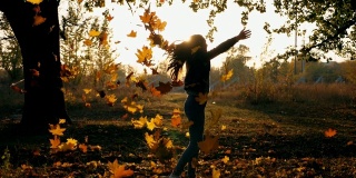 年轻的女子举起她的手，旋转着，欣喜的彩色秋叶。快乐的女孩在夕阳的背景下表现出快乐的情绪。欣赏秋色的女士。慢镜头