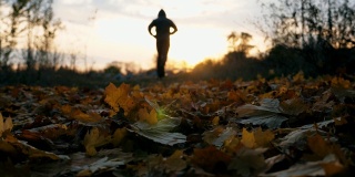 一个面目全非的运动员在秋天的公园里慢跑，踢着干枯的落叶。在日落背景下训练的男运动员的剪影。健康生活方式的概念。后视图慢动作