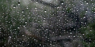 雨落在窗外绿树成荫的玻璃上。特写镜头。缓慢的运动。雨水打在玻璃上。雨滴落在车窗背景上。