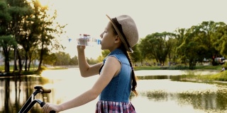 一个小女孩戴着帽子在城市公园的河边喝水，在夏天的夕阳下，坐在一辆自行车上。肖像。的处理。4 k。
