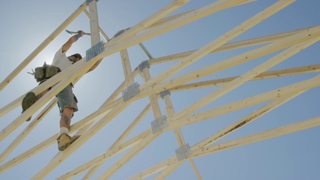 一名40多岁的白人男性建筑工人在一个晴朗、阳光明媚的日子里，用锤子和钉子将木质屋顶桁架和屋梁固定在一起