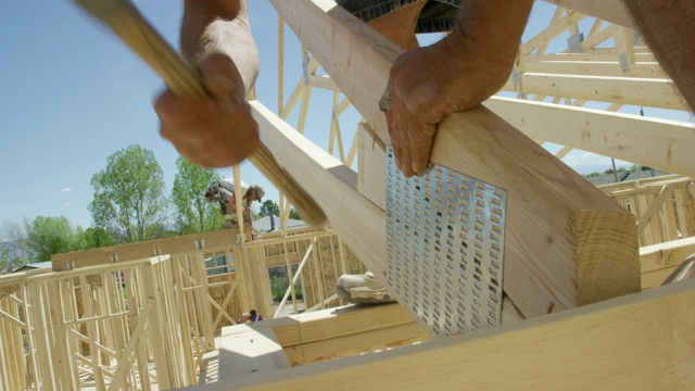 两名40多岁的白人男性建筑工人用锤子和钉子将一个木制的屋顶桁架固定在一个结构上，同时在一个晴朗、阳光明媚的日子里搭建房子并吸烟