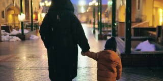 孩子和妈妈走在下雪的夜晚城市街道上。