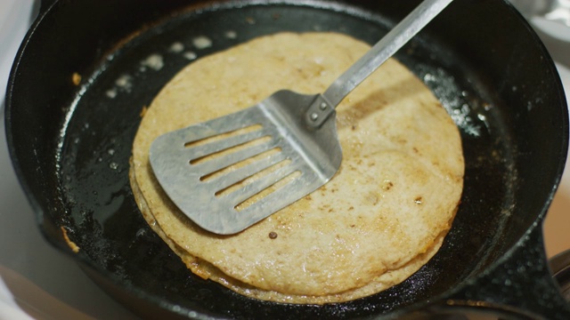 一个人用一个金属烙饼翻转在一个铸铁平底锅在炉灶上烹饪玉米粉饼(玉米饼和奶酪)