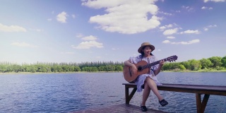 快乐的年轻亚洲女人在湖边和蓝天的背景，复制空间演奏古典吉他