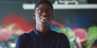 年轻黑人男性创造性地对着镜头微笑，在休闲办公室的壁画前，头和肩膀