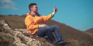 一名身穿黄色夹克、蓝色牛仔裤、戴着眼镜的男子坐在山上，欣赏风景，用手机拍摄视频。